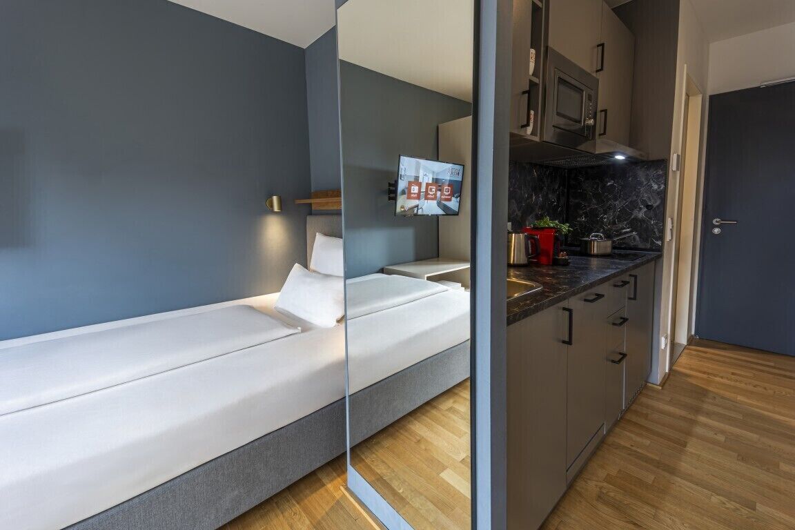 A modern room with a bed, a TV mounted on the wall, a kitchen area with cabinets and a countertop, and a wooden floor.