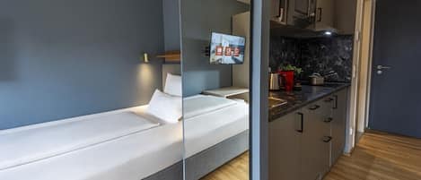 A modern room with a bed, a TV mounted on the wall, a kitchen area with cabinets and a countertop, and a wooden floor.