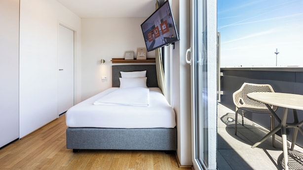 Quarto de hotel moderno com cama, televisão fixada na parede, mesa pequena e uma cadeira.
