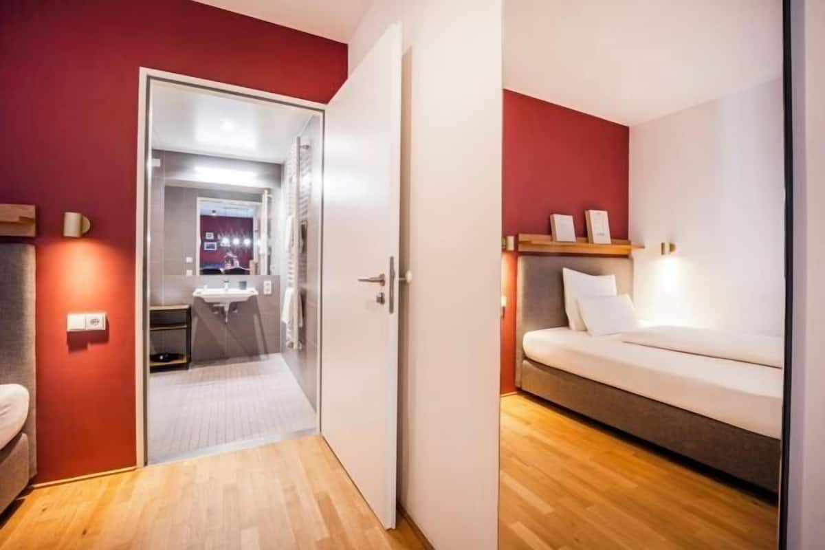 Habitación de hotel con suelo de madera, una cama, una mesita de noche, un espejo y una pared roja.