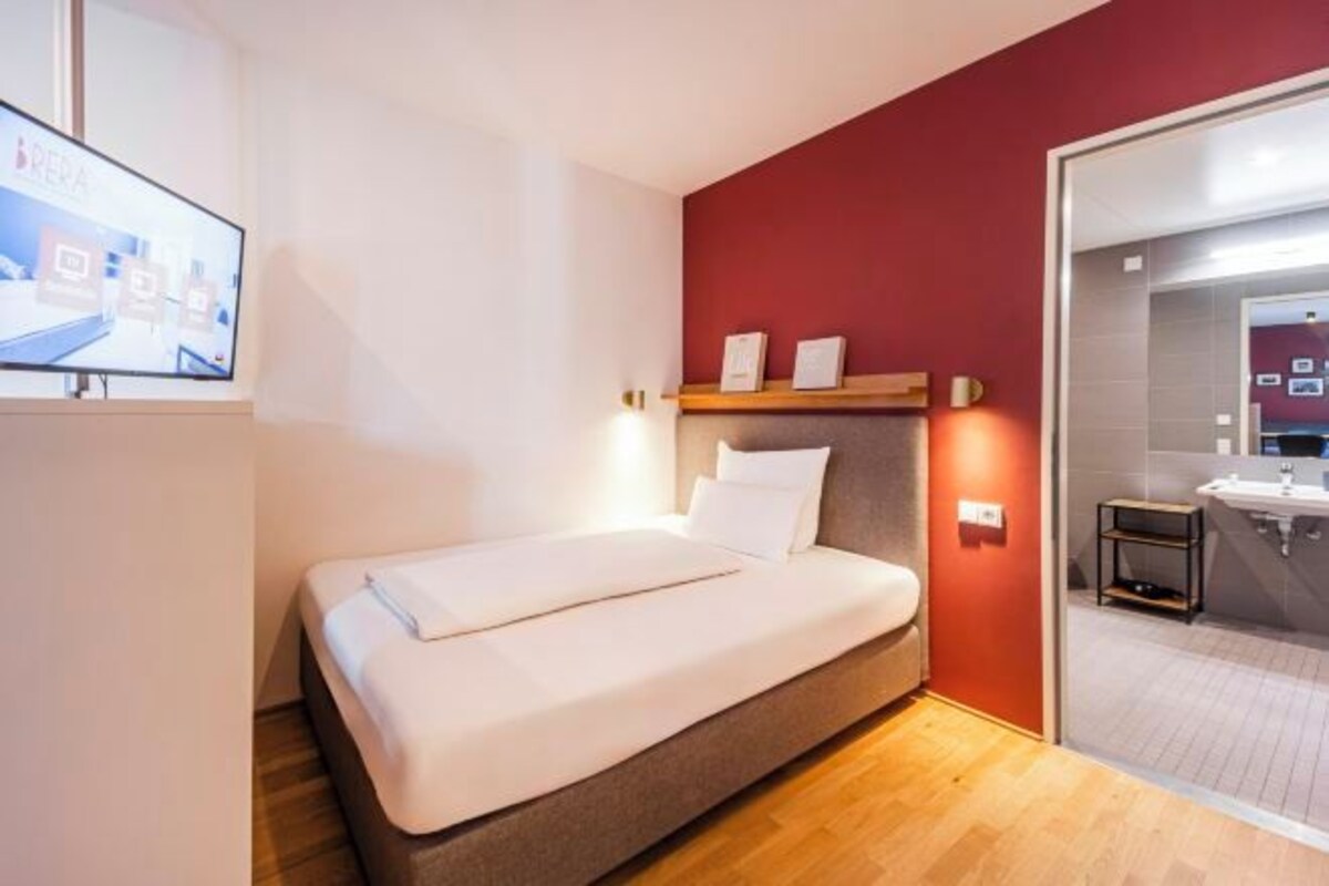 Een eenpersoonsbed in een kamer met een houten vloer, een rode accentmuur, een televisie aan de linkerkant en een zichtbare badkamer door een glazen deur.