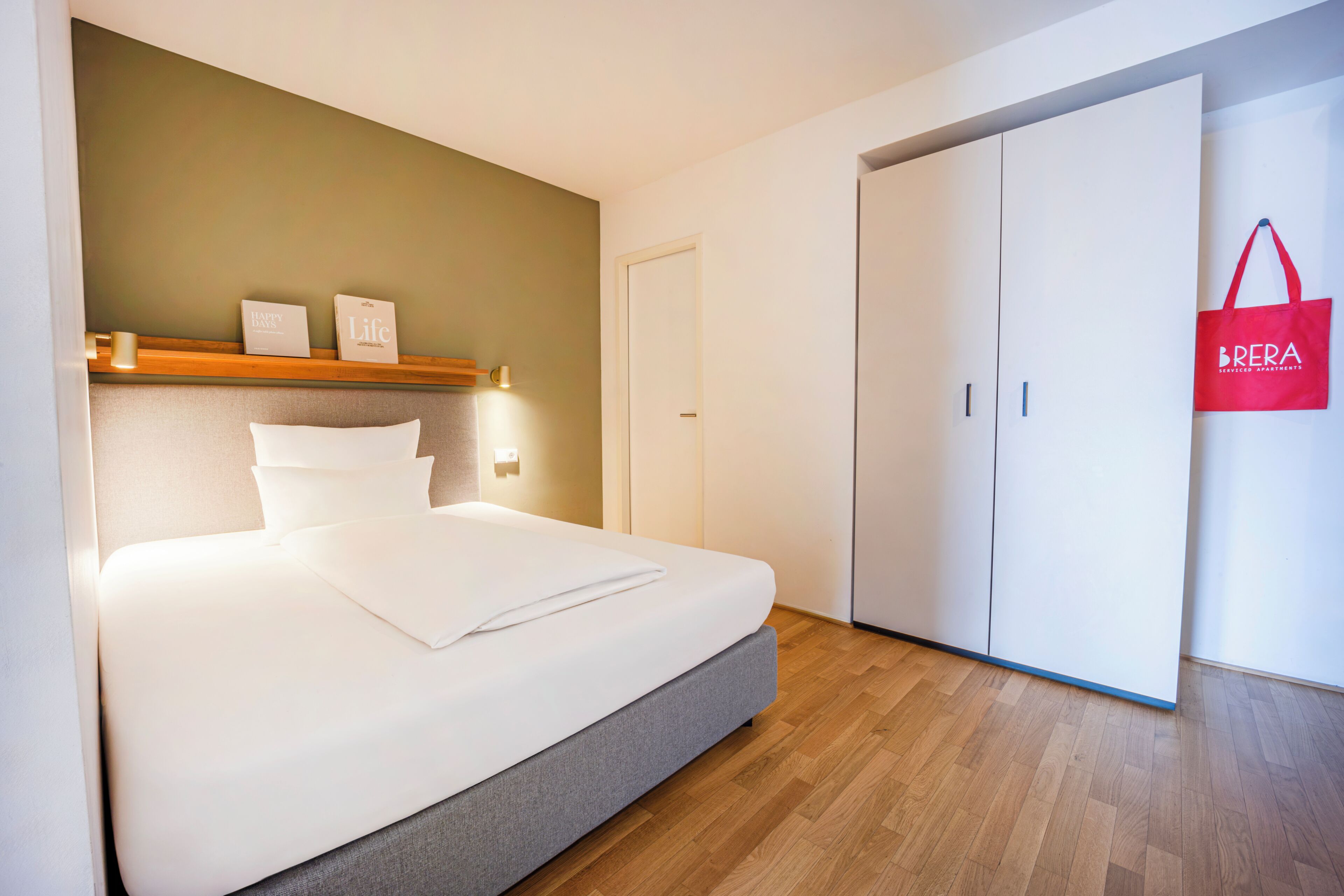 A neatly made bed with a grey headboard, a wooden shelf with two framed items, a white wardrobe, and a red bag hanging on the wall.