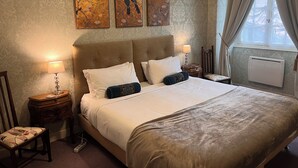 Een net opgemaakt bed met witte lakens, twee bedlampjes, een houten nachtkastje met lamp, een stoel met een gestreept kussen, en drie ingelijste kunstwerken aan de muur.