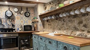 Een keuken met een houten aanrecht, een fornuis en een magnetron. Er hangen potten en pannen aan de muur en een plank met bekers en borden.