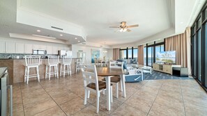 A spacious living area with a dining table, white chairs, a ceiling fan, and large windows offering a view. - Wow!! New Owner and New to Rental Market! (Orange Beach)