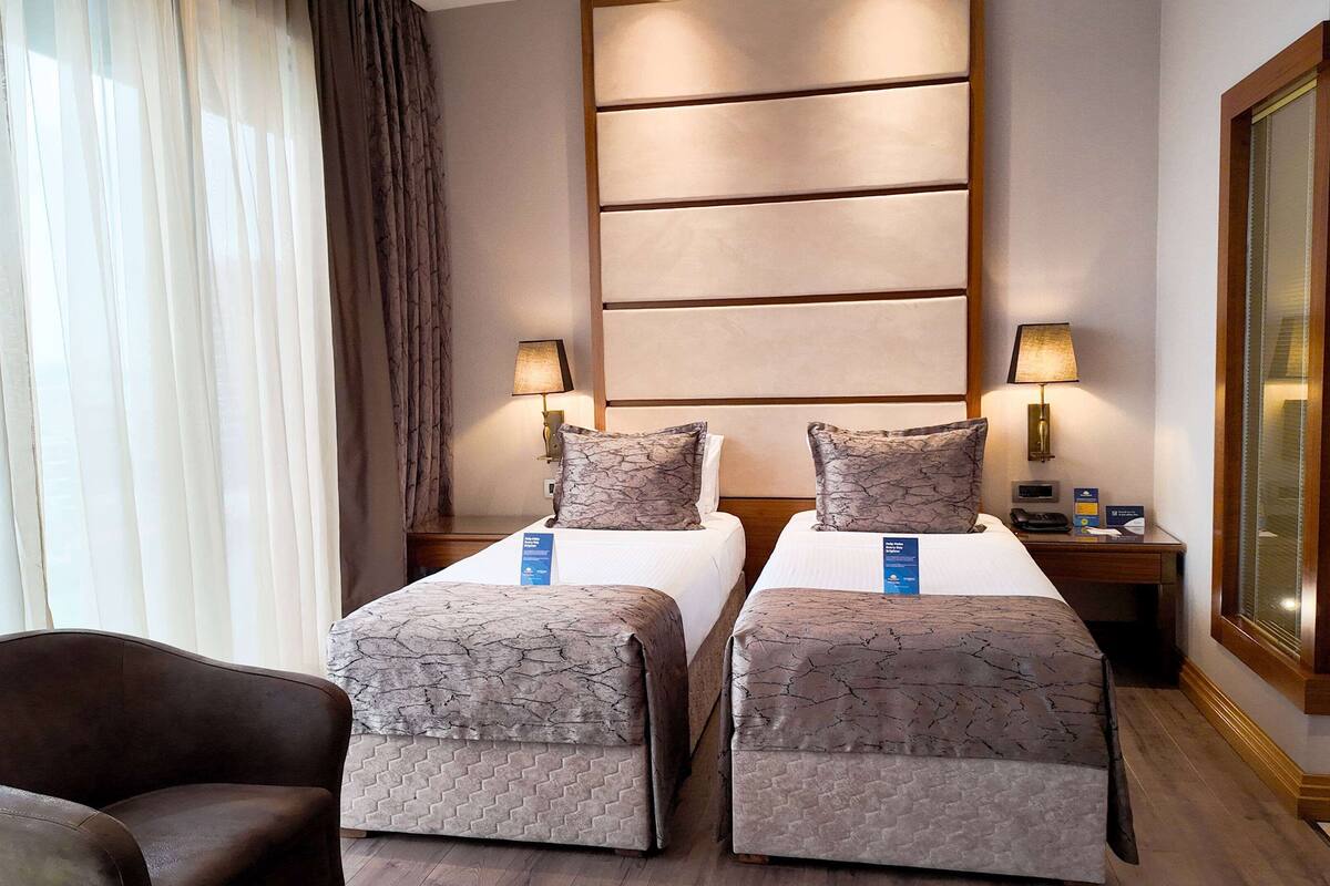 Une chambre d’hôtel avec deux lits, une table de chevet, une lampe, une chaise et une grande fenêtre avec des rideaux.