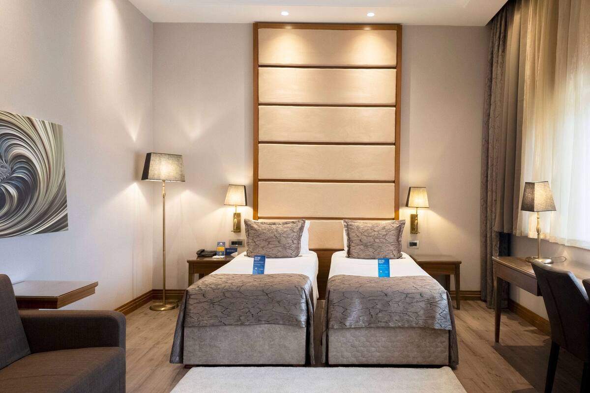 Une chambre d’hôtel avec deux lits, un bureau, une chaise et un grand tableau abstrait accroché au mur.