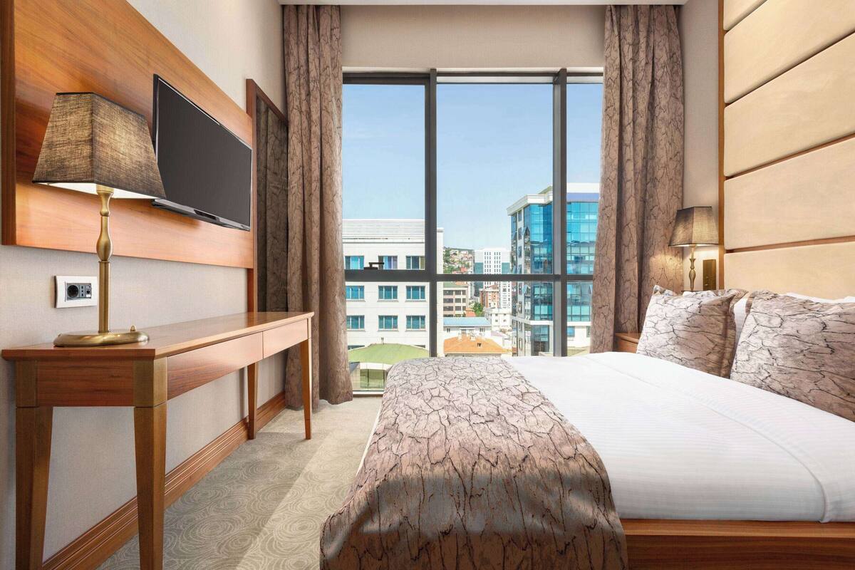 Une chambre d’hôtel dotée d’un grand lit, d’un bureau, d’un téléviseur à écran plat fixé au mur et offrant une vue sur les bâtiments à travers de grandes fenêtres.