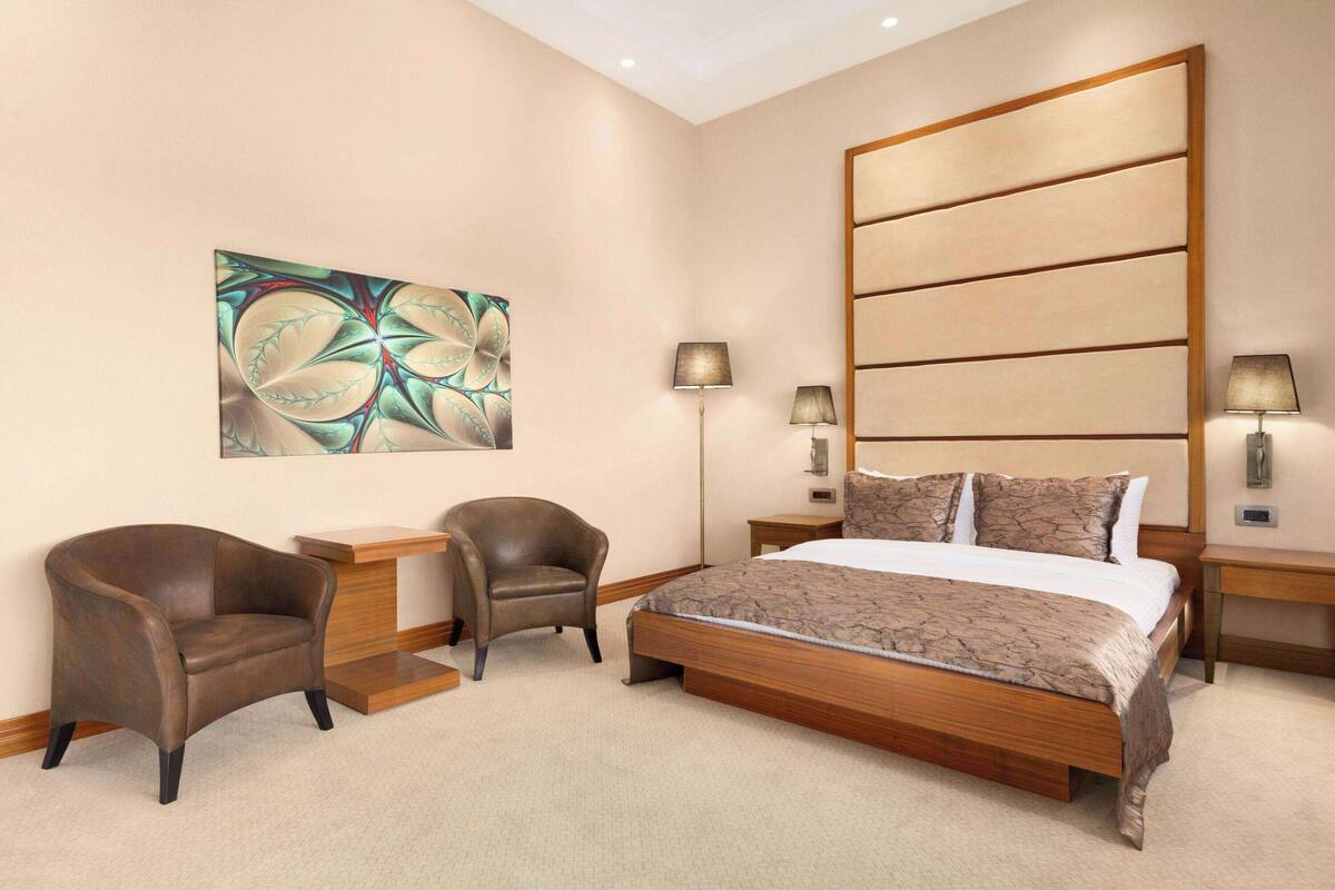 Une chambre d’hôtel moderne, dotée d’un grand lit, de deux fauteuils, d’une table d’appoint, d’une table de chevet et d’un élément décoratif mural.
