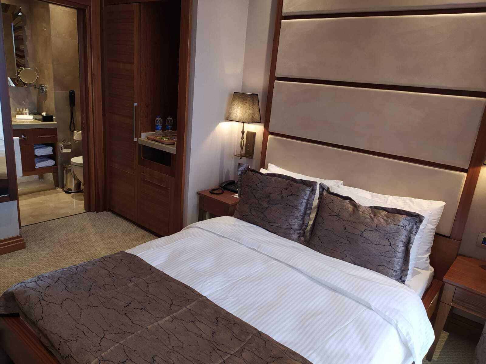 A hotel room with a bed, a nightstand, a lamp, a wooden wardrobe, and a bathroom visible through an open door.