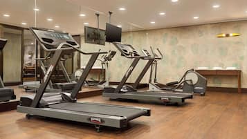 Un gimnasio con varias cintas de correr, un televisor en la pared y piso de madera.
