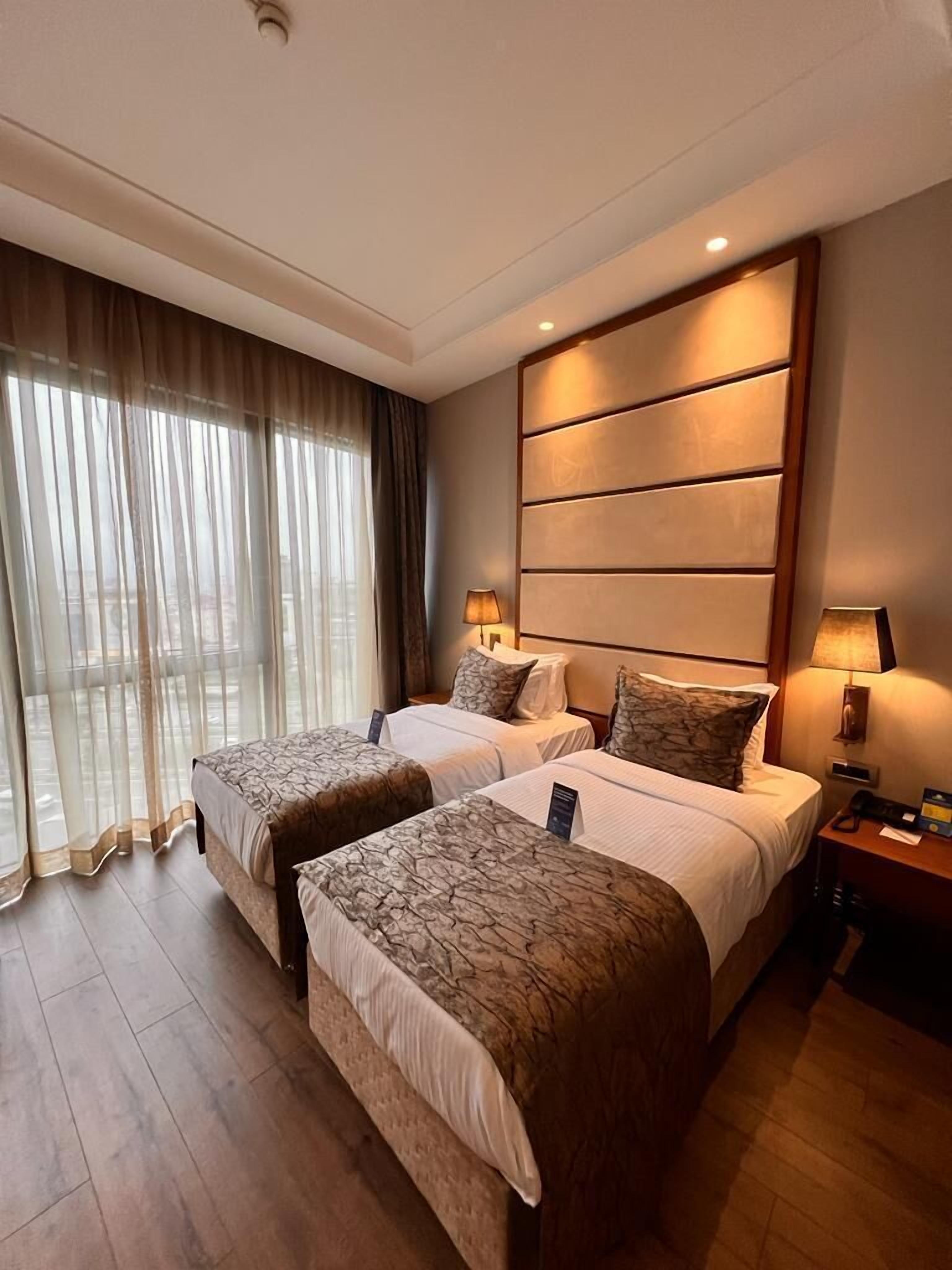 Une chambre d’hôtel avec deux lits, une grande fenêtre avec des rideaux, un panneau fixé au mur et une table de chevet avec une lampe.