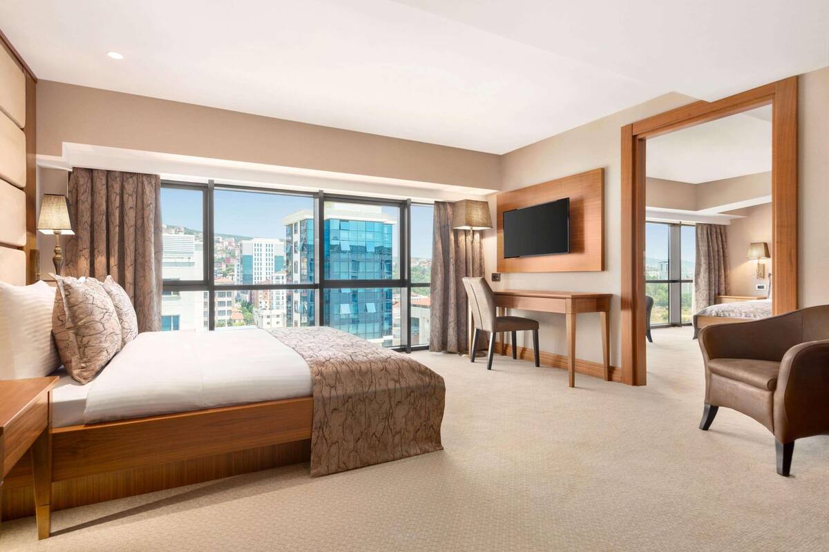 Une chambre d’hôtel équipée d’un lit, d’un bureau, d’une chaise, d’une télévision et de grandes fenêtres donnant sur la ville.