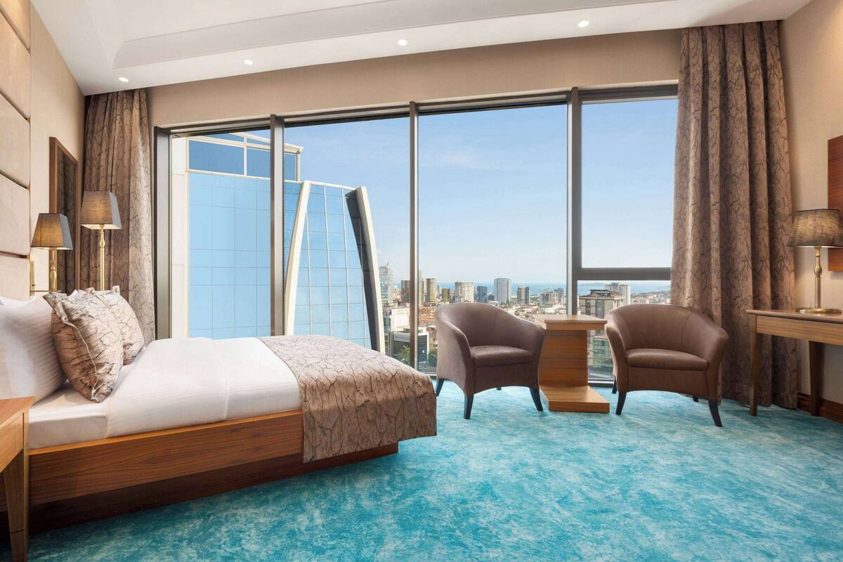 Une chambre d’hôtel moderne dotée d’un grand lit, de deux fauteuils, d’une petite table et d’une grande fenêtre donnant sur la ville.