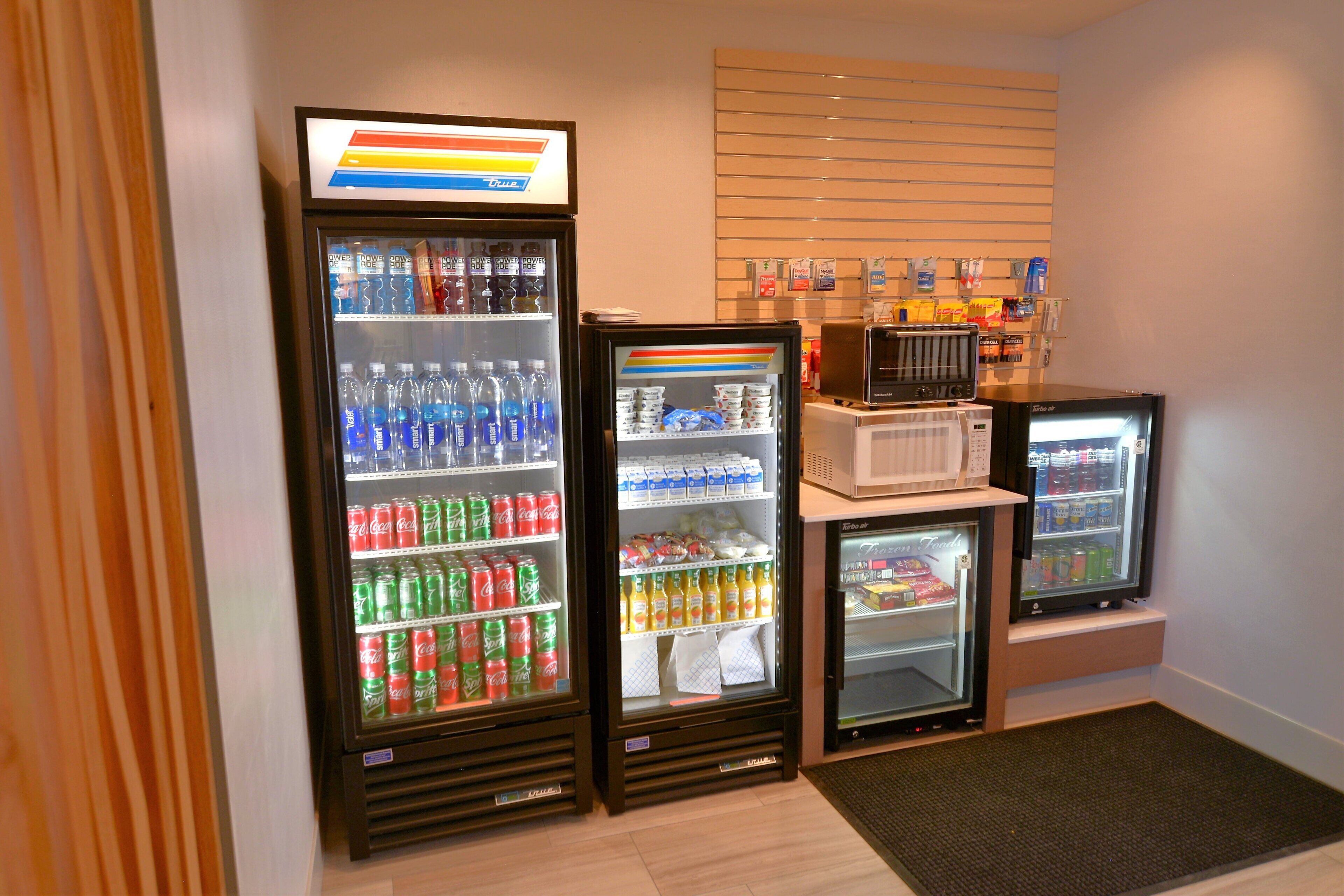 Una hilera de refrigeradores para bebidas llenos de diversas opciones, un microondas y una pequeña selección de aperitivos.