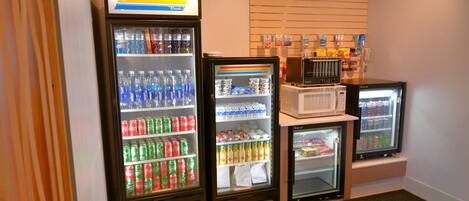 Una hilera de refrigeradores para bebidas llenos de diversas opciones, un microondas y una pequeña selección de aperitivos.