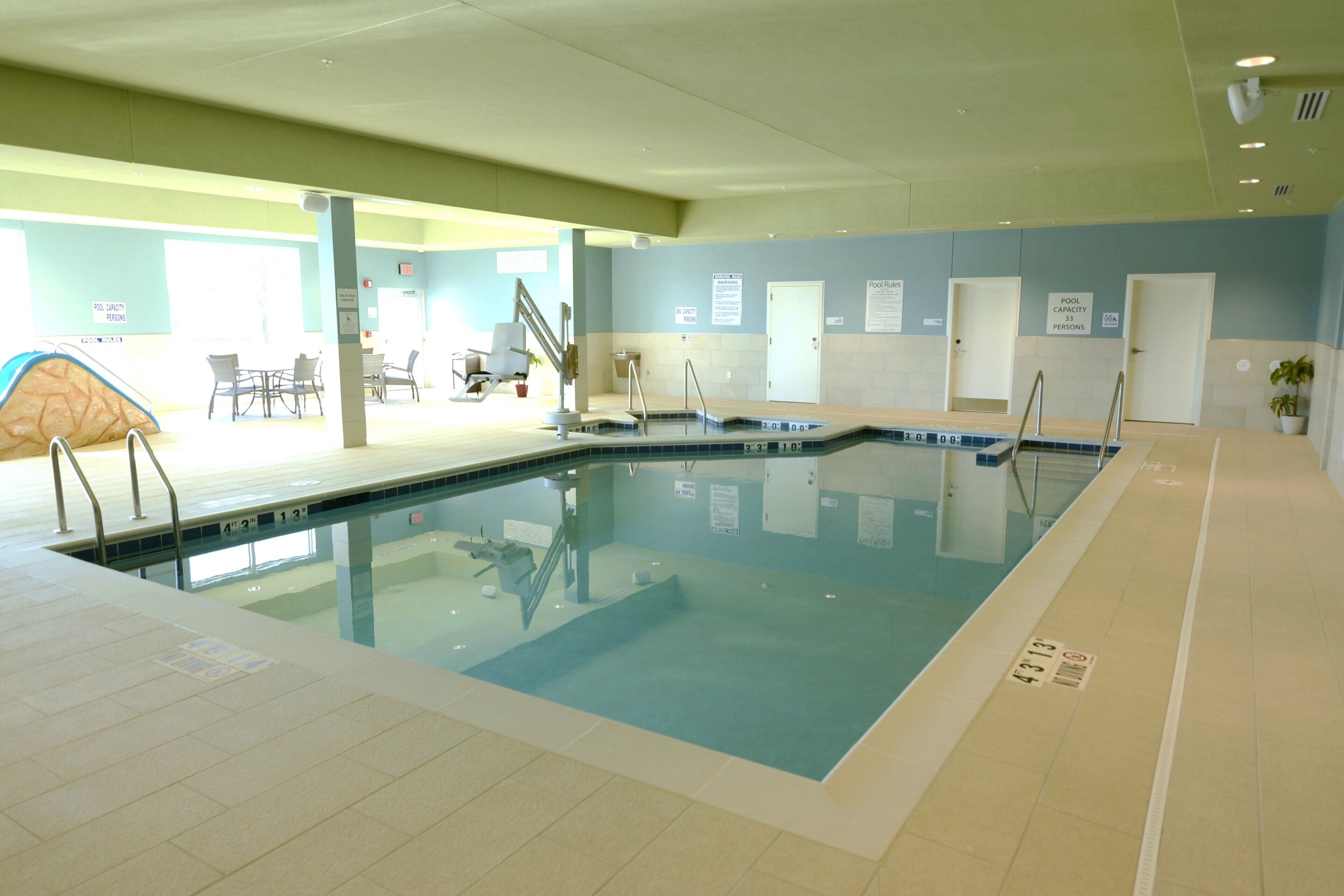 An indoor swimming pool with a lifeguard chair, pool capacity signs, and a sunshade.