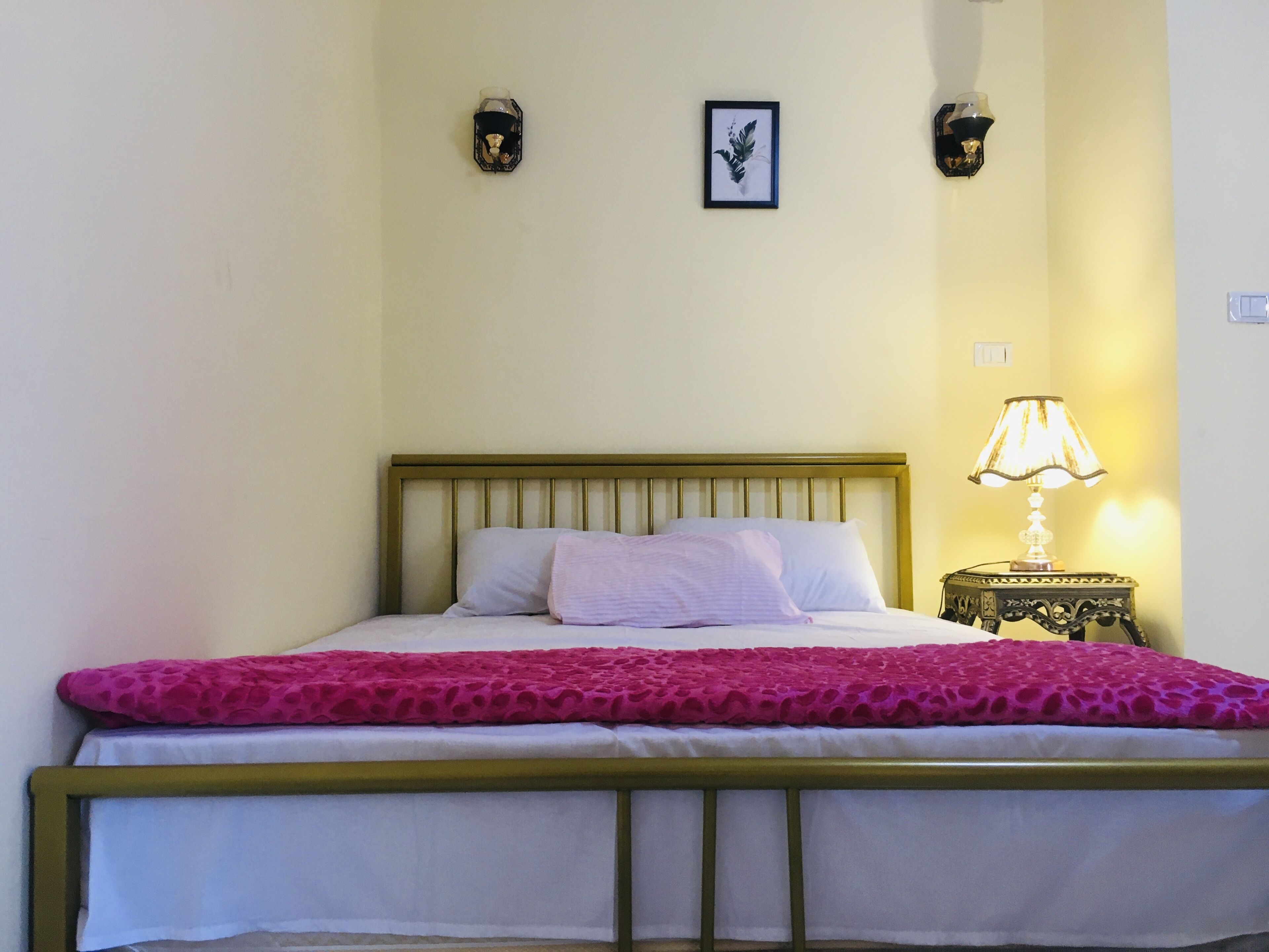 Ein Bett mit Holz-Kopfteil, weißer Bettwäsche und einer rosa, strukturierten Decke. Ein Nachttisch mit einer Lampe und ein gerahmtes Bild an der Wand sind ebenfalls zu sehen.
