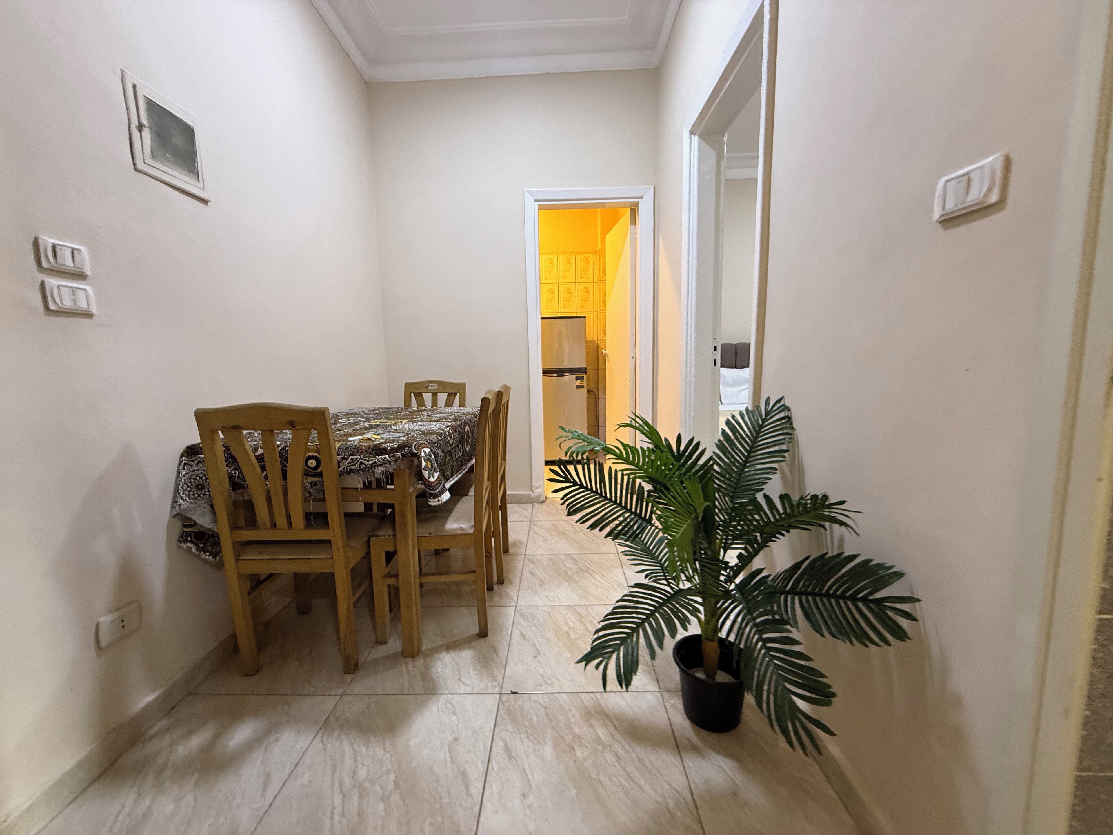 Un couloir étroit avec une table et des chaises, une plante en pot et une porte donnant sur la cuisine.