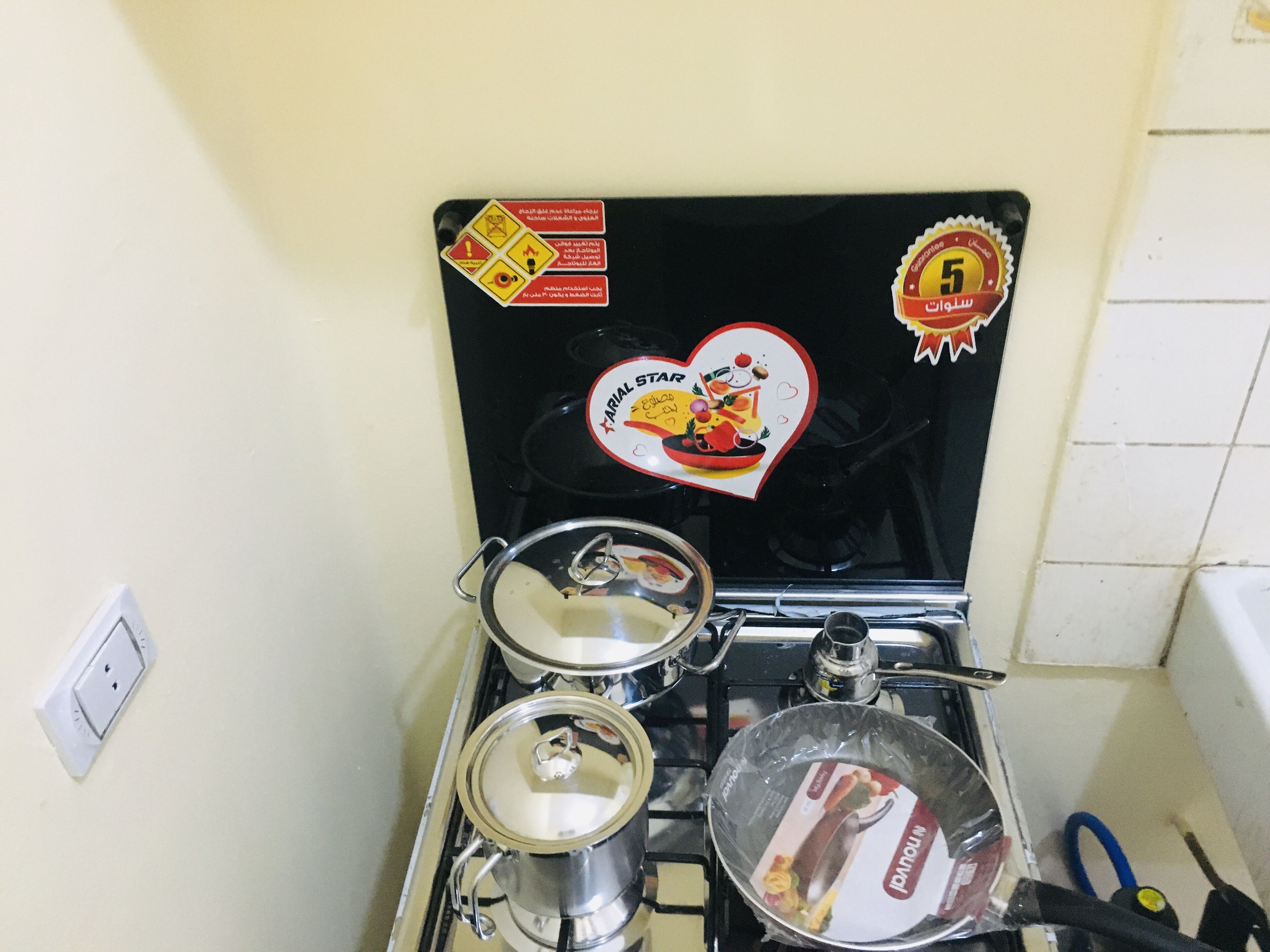A kitchen with a stove, pots, and a pan. There are stickers on the stove, including a heart-shaped one with a food illustration.