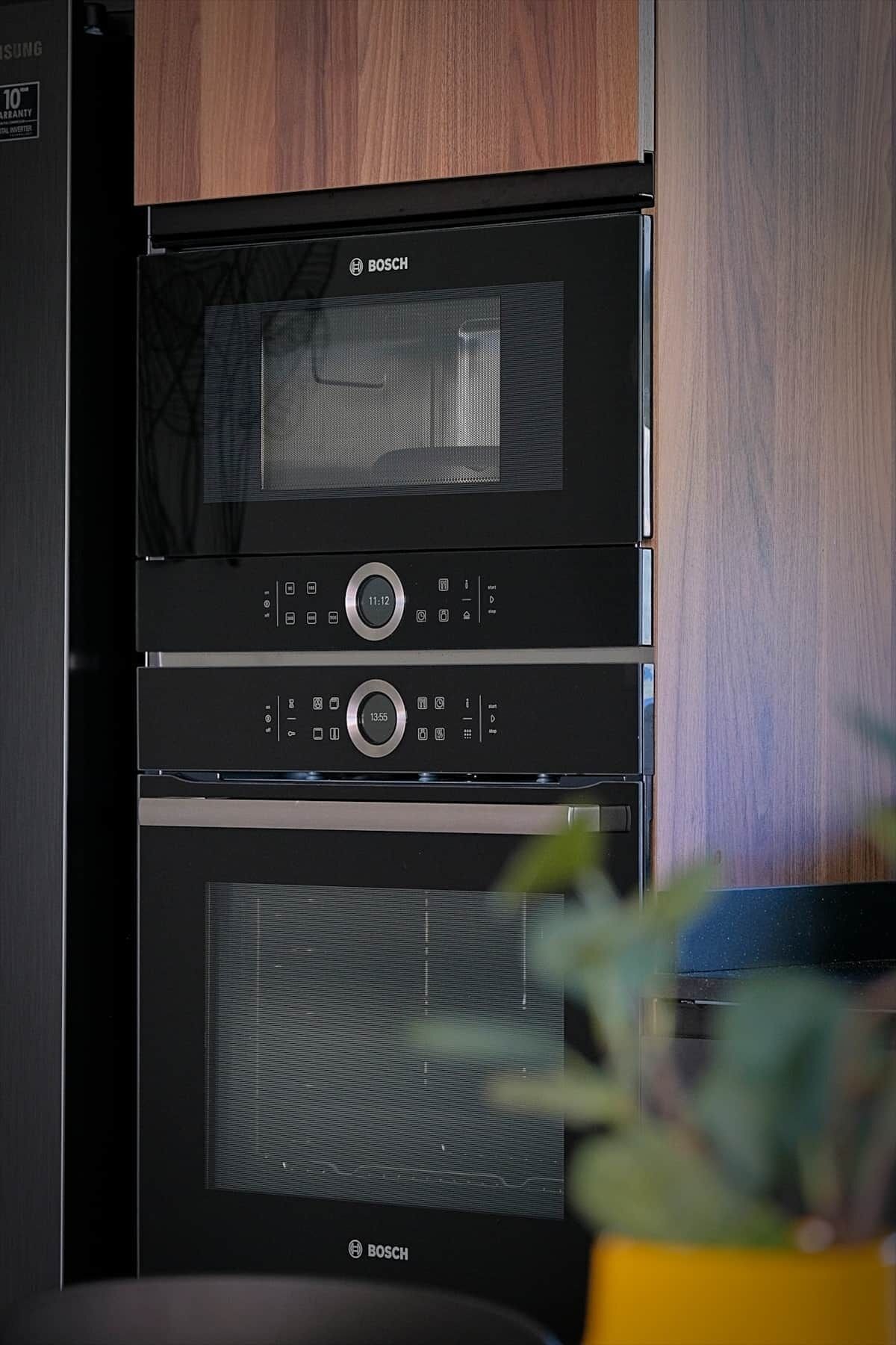 A Bosch oven with a digital clock display, a microwave above it, and a plant in the foreground.