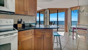 A kitchen with wooden cabinets, a white stove, and a black countertop. There is a dining area with a table and chairs, and a view of the ocean through large windows.