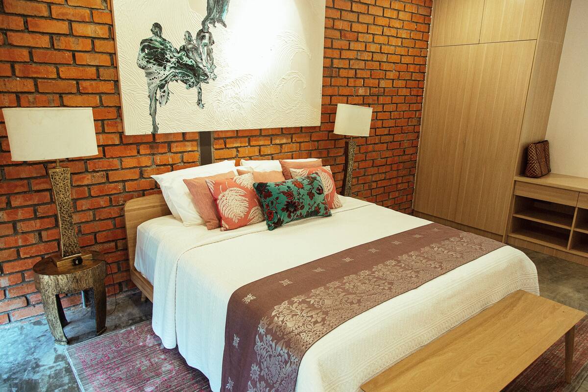 Een slaapkamer met een bakstenen muur, een bed met veel kussens, een houten bankje en een schilderij boven het bed.
