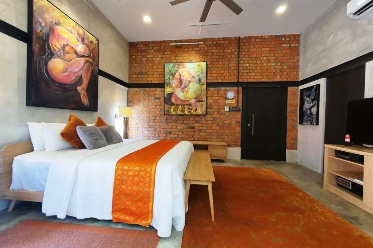 Habitación de hotel con pared de ladrillo, cama con ropa de cama blanca y detalles naranja, escritorio, silla, televisor y obras de arte en las paredes.
