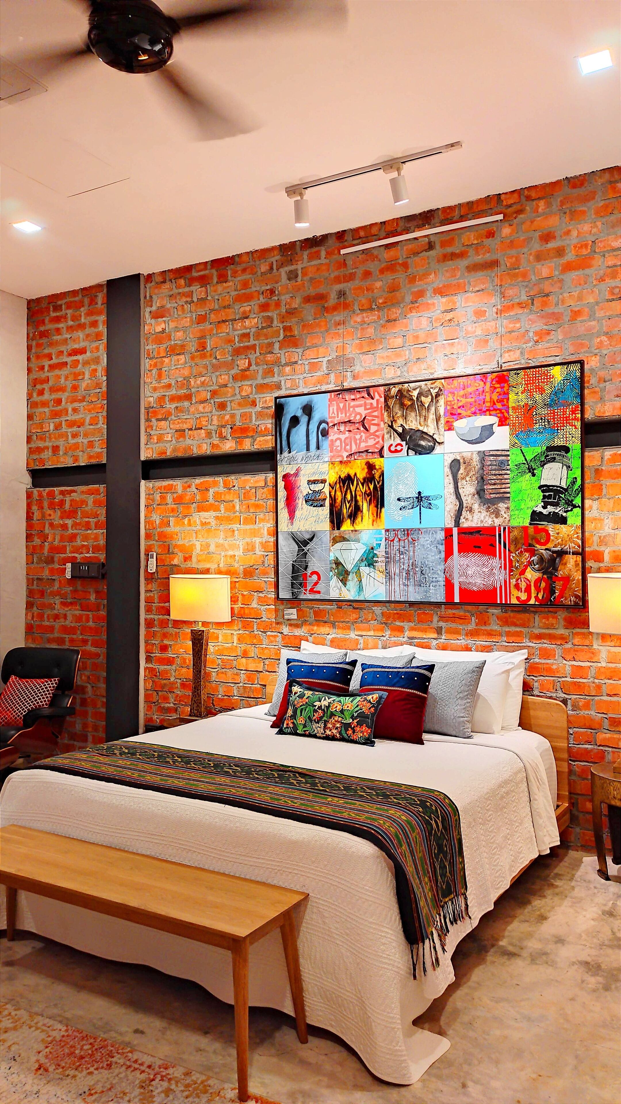 Un dormitorio con pared de ladrillo, una gran obra de arte abstracto, cama con muchas almohadas y ventilador de techo.