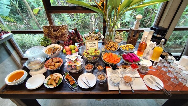 A buffet table with various dishes and drinks, set against a backdrop of lush greenery visible through a window.