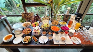 Una mesa de buffet con diversos platos y bebidas, con un fondo de vegetación exuberante visible a través de una ventana.