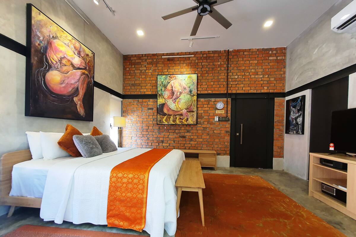 Een moderne hotelkamer met een bakstenen muur, een groot bed met witte beddengoed en oranje accenten, een houten bankje, een televisie en kunstwerken aan de muren.