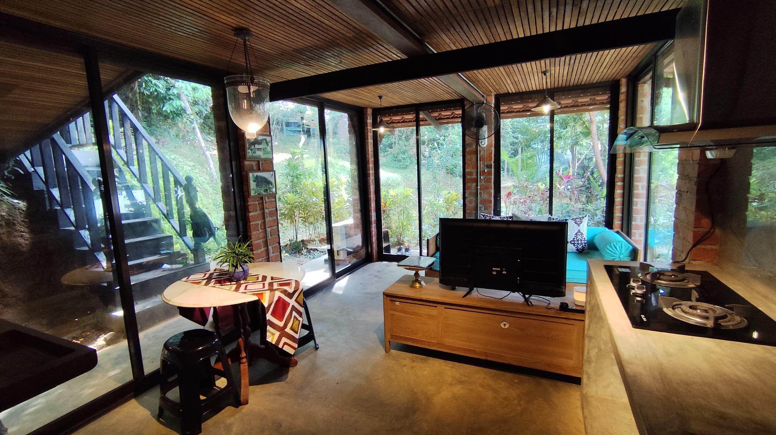 A modern living room with a TV, wooden furniture, and large windows offering a view of a staircase and greenery.
