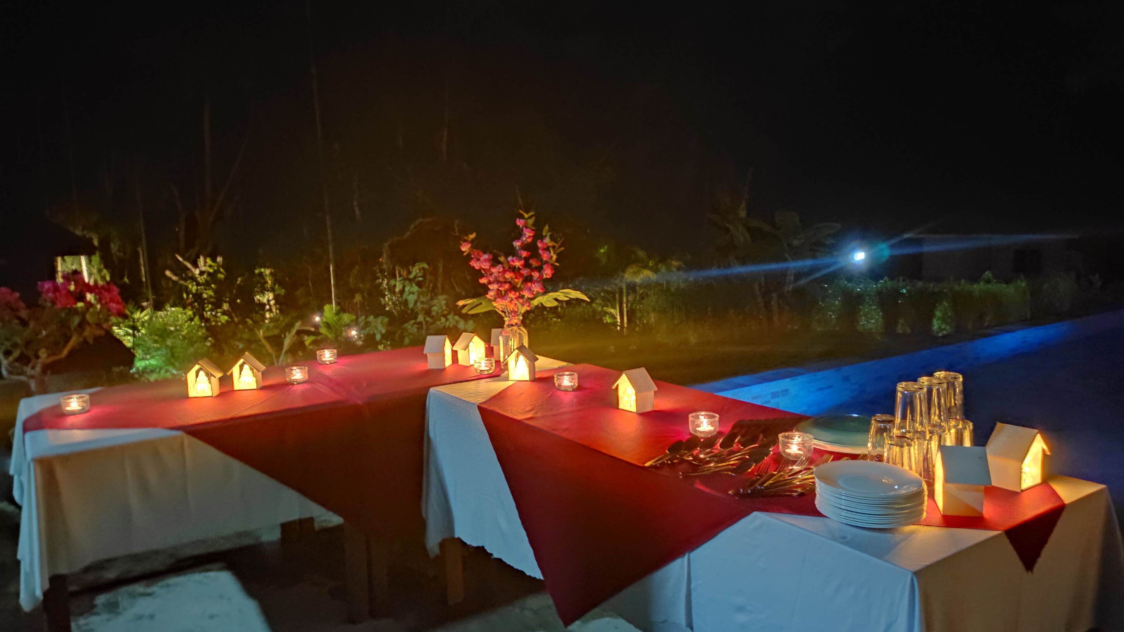 An outdoor dining setup with tables covered in red tablecloths, lit by candles and small house-shaped lights.