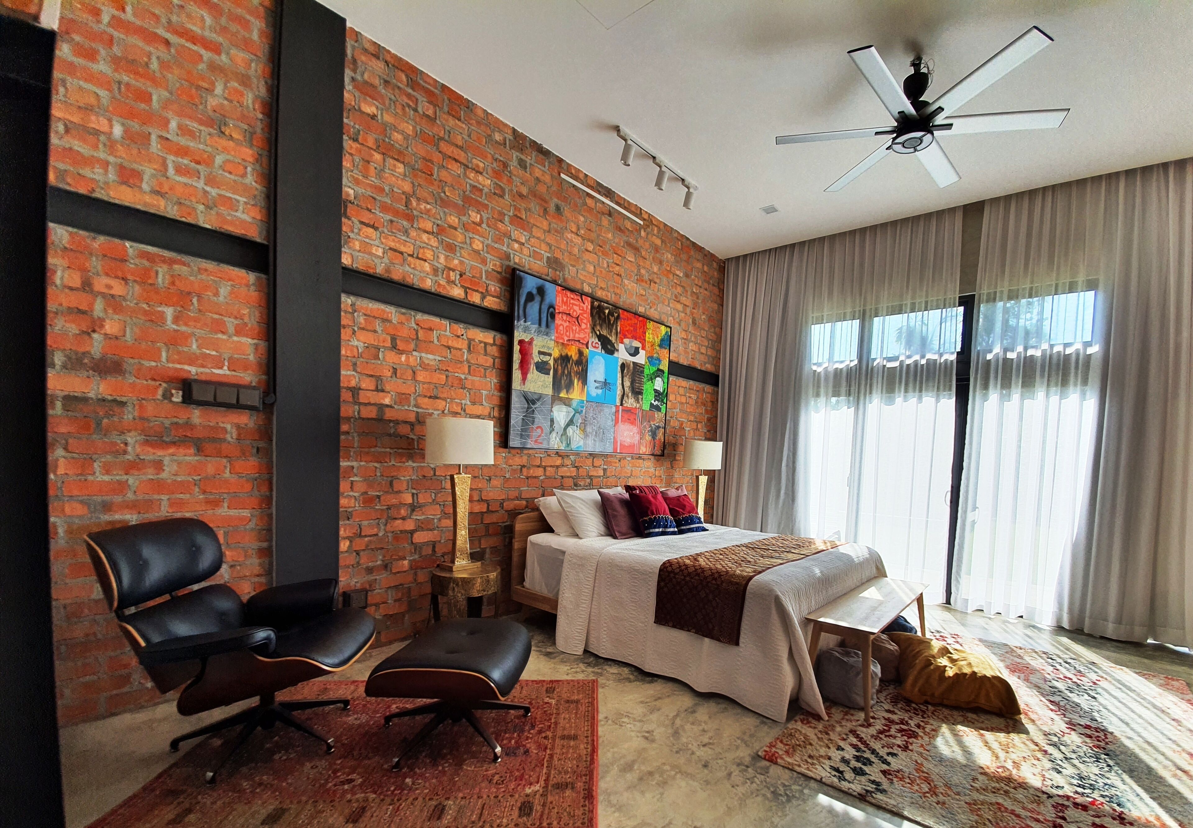 Ein Hotelzimmer mit Backsteinwand, einem Bett mit weißer Tagesdecke, einem Schreibtisch, einem Stuhl und einem großen Fenster mit Vorhängen.