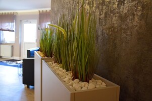 Ein modernes Interieur mit einer langen Pflanzkasten-Anlage, die hohe, grüne Pflanzen und weiße Kieselsteine enthält, einem blauen Sofa und einer strukturierten Wand.