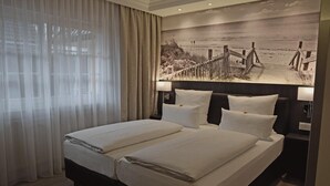A double bed with white linens, a headboard with a painting of a beach scene, and a bedside table with a lamp.