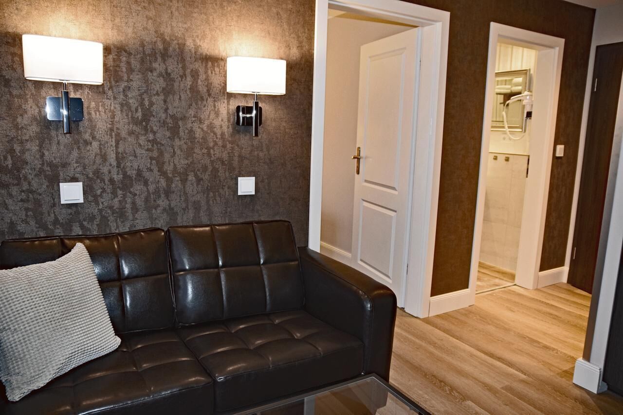 A modern living room with a dark leather sofa, two wall-mounted lamps, and a wooden floor.