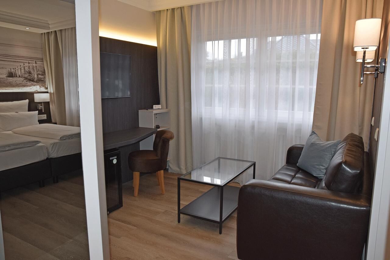 Een hotelkamer met een bed, een bureau, een glazen bijzettafeltje, een leren bank en een groot raam met transparante gordijnen.