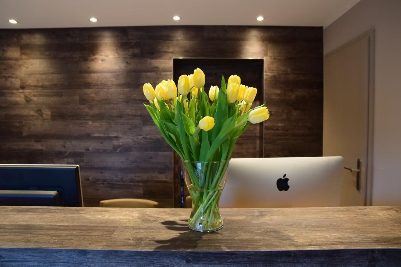 A wooden table with a clear glass vase containing yellow tulips, an Apple laptop, and a dark wooden wall in the background.
