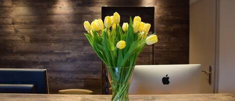 A wooden table with a clear glass vase containing yellow tulips, an Apple laptop, and a dark wooden wall in the background.