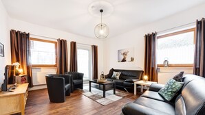 A modern living room with a sofa, two armchairs, a coffee table, and a side table. There is a window with curtains, a wall-mounted picture, and a ceiling light fixture.