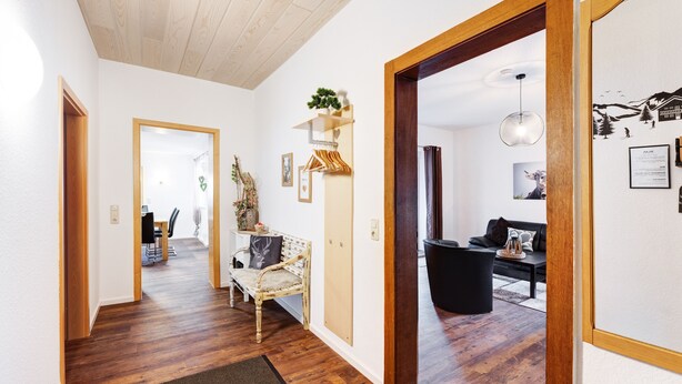 A hallway with wooden flooring, a bench, a black trash bin, and a wall with a framed picture and a small plant.