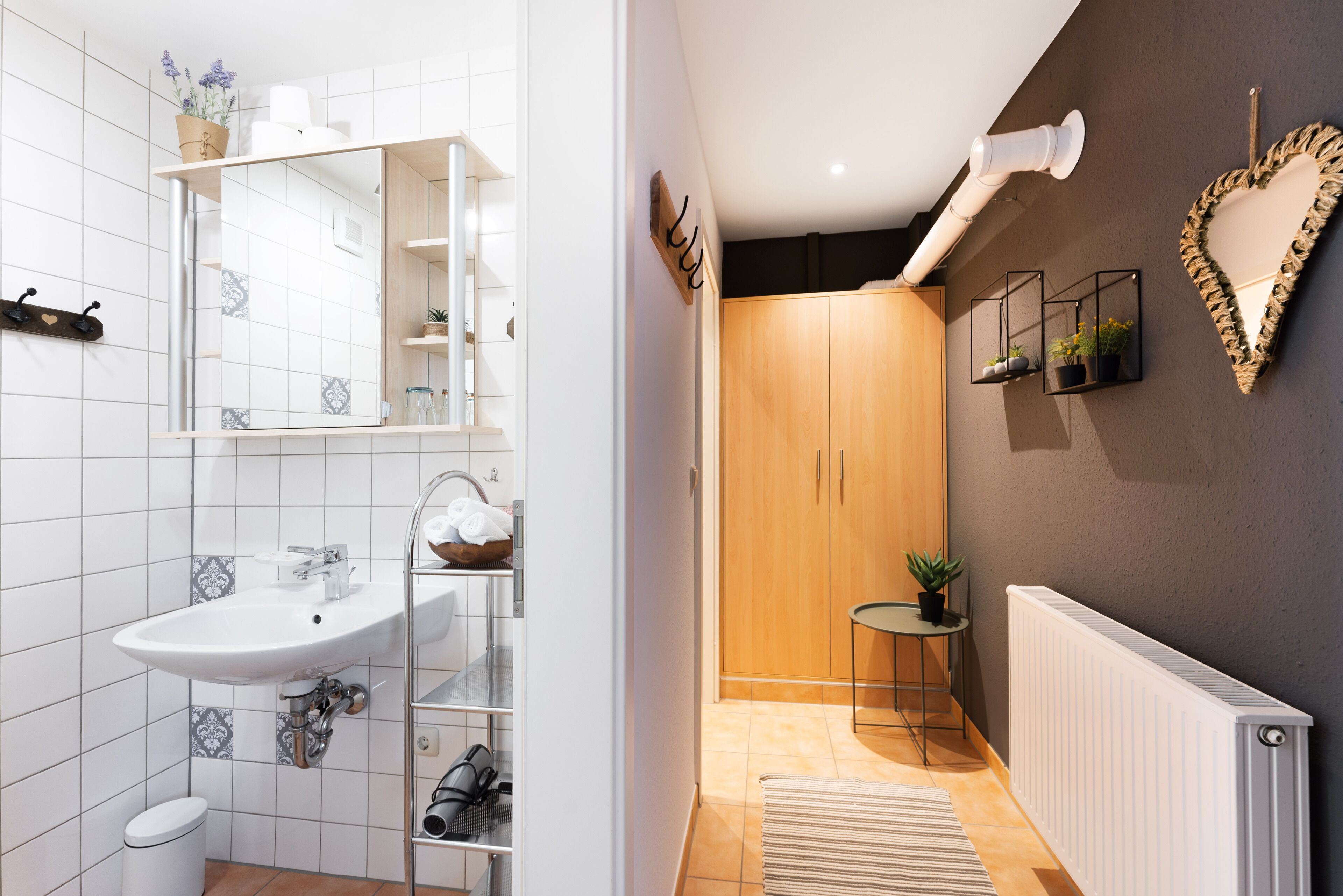 Un bagno moderno con lavandino bianco, specchio e pareti piastrellate. Si nota un mobiletto in legno e un tavolino con una pianta.