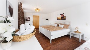 A bedroom with a white bed, wooden flooring, a bedside table, a wall-mounted light fixture, and a decorative wall piece.