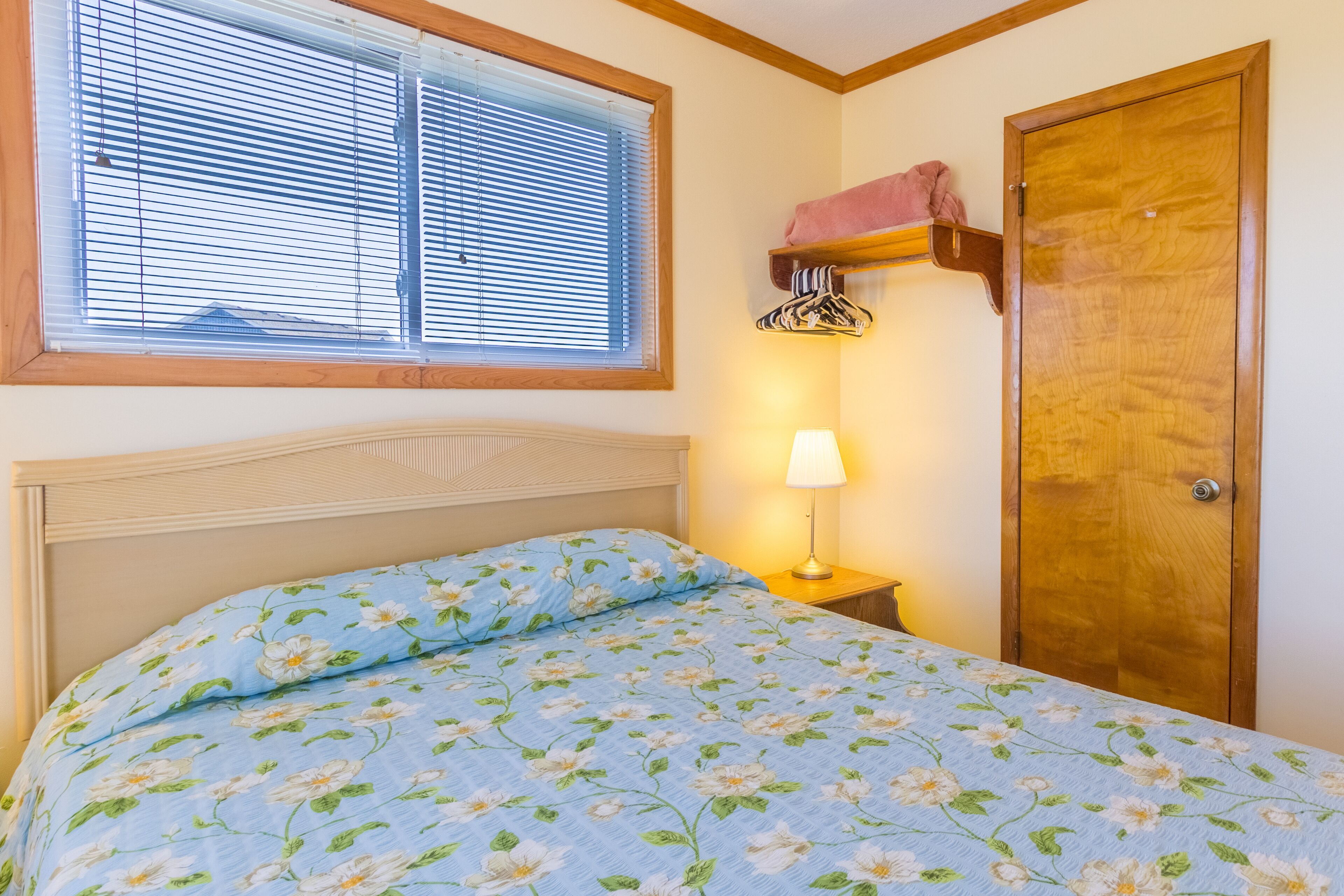 A bedroom with a bed, a nightstand, a lamp, a window with blinds, and a wooden door.