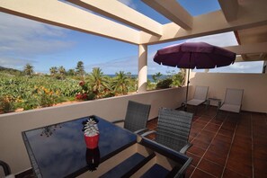 Eine Terrasse mit Tisch und Stühlen, einem violetten Sonnenschirm und Blick auf das Meer und die Vegetation.