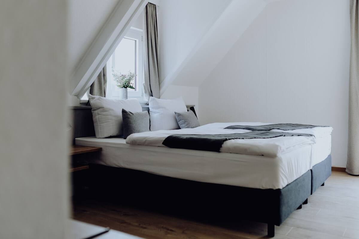Un dormitorio con cama, almohadas, ventana y cortinas.