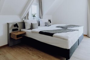 Un dormitorio moderno con techo inclinado, cama con ropa de cama blanca y detalles en gris, mesita de noche con lámpara y ventana con cortinas.