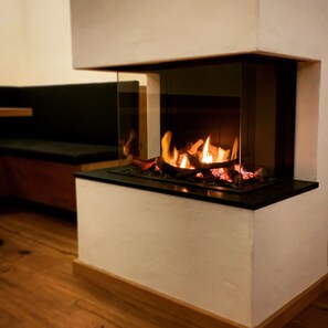 Una chimenea moderna con frente de vidrio, ubicada en un ambiente minimalista con piso de madera y un banco de color oscuro.